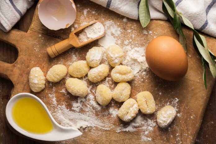 Ingredientes para preparar gnocchi caseiro, incluindo ovos, farinha e azeite, em uma tábua de madeira.